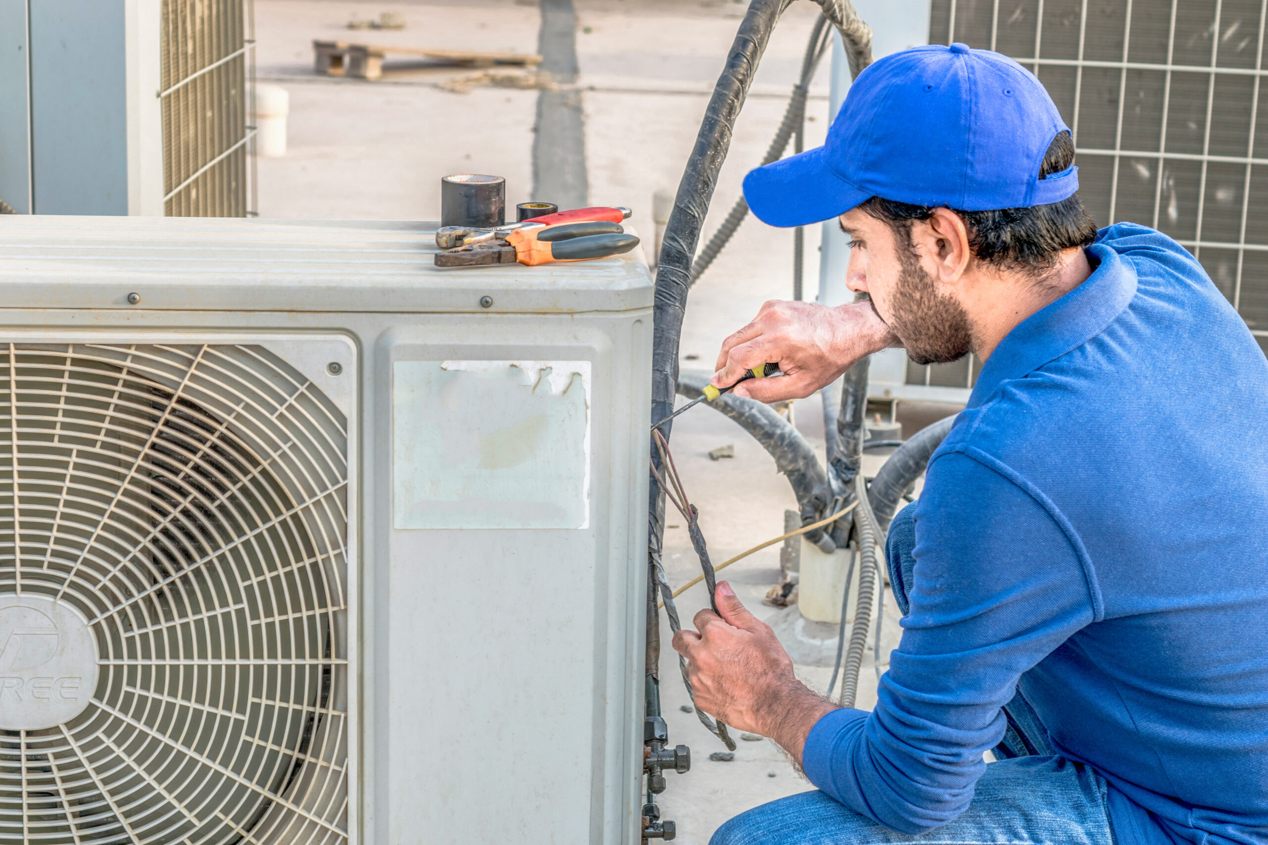 a professional electrician man is fixing a heavy duty unit of central air conditioning system by his tools on the roof top and wearing blue color of uniform and white cap